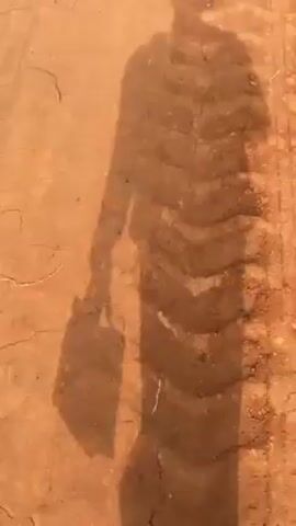 The video shows a person walking along a reddish-brown dirt path. The person is wearing sandals and their shadow is visible on the ground. The shadow appears to be holding a rectangular object. The path is cracked and uneven, with some small rocks and debris along the edge.