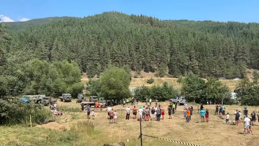 The video shows a wide shot of an outdoor event with a large gathering of people and vehicles in a grassy, hilly area. The background is dominated by a dense, green forest covering a mountain slope under a clear blue sky. In the foreground, a dry, grassy field is filled with numerous people, many standing in clusters and some seated. Several off-road vehicles, including trucks and jeeps, are parked around the perimeter of the gathering and in the middle of the field. Some of the vehicles appear to be modified or rugged, suggesting an off-road event. There are also tents or canopies set up in the distance, possibly for vendors or spectators. Yellow and black caution tape is visible in the lower right corner of the frame, marking a boundary. The overall atmosphere appears to be a casual outdoor event, likely an off-road competition or gathering, on a sunny day.