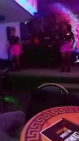 Here is a concise description of the video:

The video shows two women dancing on a stage in what appears to be a nightclub or outdoor lounge. They are wearing black tops and bright pink shorts. There is a DJ booth behind them. The camera is positioned at a table with a menu. There are also other tables and chairs in the foreground. The lighting in the establishment is dark, with colored lights illuminating the stage and the dancers. The overall atmosphere suggests a party or entertainment setting.