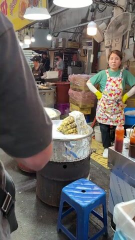 The video shows a bustling outdoor market scene. In the foreground, two large metal drums serve as steaming stations, each topped with a perforated metal tray. The tray on the left holds several ears of corn covered in plastic wrap, appearing white and steamed. The tray on the right displays ears of corn with alternating yellow and brown kernels, also covered in plastic. A woman in a floral apron and yellow gloves stands behind the steaming stations, facing the camera with her hands on her hips. Behind her, a food stall is visible with several people preparing food and customers interacting. The stall is equipped with metal counters, cooking equipment, and stacks of boxed goods. The environment suggests a lively food market with various vendors and customers.