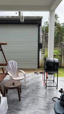 A shirtless man wearing swim trunks and a baseball cap walks from the left side of the frame towards a large, ribbed metal roll-up door at the back of a porch. He is holding his right hand up as if to indicate something or to signal a command. The porch has a tiled floor and is furnished with a wooden swing, a white plastic chair with a towel draped over it, and a small brown plastic table. To the right of the door, there is a black grill on a stand. The man approaches the door and appears to be interacting with it, though his exact action is unclear in this short clip. The overall environment appears to be outdoors, possibly a backyard or patio area, with some greenery visible in the background.