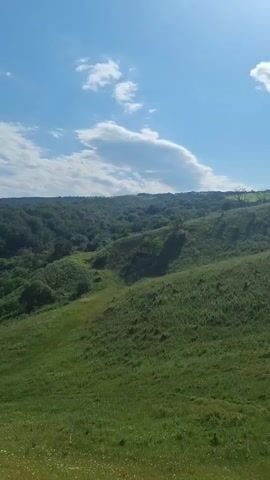 The video shows a rolling green landscape under a bright blue sky with scattered clouds. The foreground is a grassy hill with some wildflowers. In the midground, there are more hills covered in dense green trees. In the distance, there are layers of mountains. The sun is shining, creating shadows on the hills. The camera pans slowly to the right, revealing more of the expansive natural scenery.