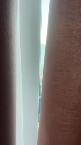 The video pans from closed curtains to reveal a balcony overlooking a beach. The beach is pebbly with rows of white lounge chairs. The sea is calm and stretches to the horizon under a clear sky.