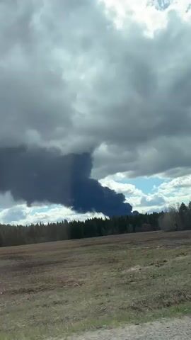 A video taken from a moving vehicle shows a large, dense column of black smoke rising into the sky from behind a line of trees, suggesting a significant fire. The smoke plume extends high into an overcast sky, which also features patches of blue and lighter clouds. In the foreground, a barren field with visible furrows stretches across the frame, bordered by a strip of dry, sparse vegetation and some leafless trees closer to the camera. A lush green forest forms the backdrop where the smoke originates. The scene is located in or near Башкултаево, Russia. Music is audible in the background.