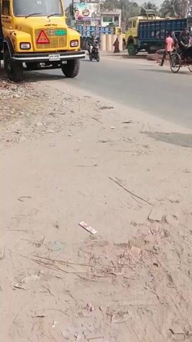 The video shows a yellow truck parked on the side of a paved road. The truck has a blue, green, and red decorated roof and a triangular sign on the front. To the left of the truck is a dirty shoulder with trash. A man wearing a suit walks across the road in front of the truck. There is a scooter and other trucks further down the road. The scene appears to be in a rural or semi-urban environment.
