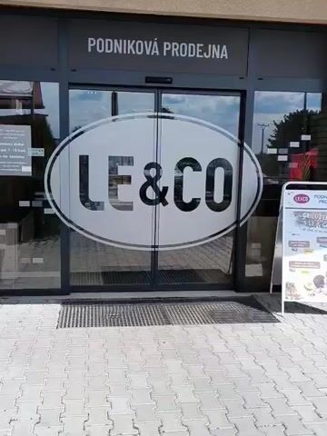 The video displays the exterior of a "LE&CO PODNIKOVÁ PRODEJNA" (LE&CO Company Store/Factory Outlet) in Jesenice, Czechia, on a bright, sunny day.

The primary subject is the store's entrance, featuring automatic glass sliding doors. A large, white oval logo with "LE&CO" in black letters is prominently displayed on the doors. Above the entrance, the text "PODNIKOVÁ PRODEJNA" is visible in white lettering against a dark background.

To the right of the doors, a white A-frame sign advertises "GRILOVÁNÍ S LE&CO" (Grilling with LE&CO), showing images of various meat products and their prices. On the left side of the entrance, a concrete trash can with some litter is present.

As the video progresses, the automatic doors slide open, revealing a white sign affixed to the interior of the left door panel. This sign provides detailed information including the company name ("LE & CO - Ing. Jiří Lenc, s.r.o."), address ("Na Orlovech 403, 252 42 Jesenice u Prahy"), and operating hours: "po-pá 7-19 hod. so-ne zavřeno" (Monday-Friday 7 AM - 7 PM, Saturday-Sunday closed). A website address ("www.masoleco.cz") and email ("info@masoleco.cz") are also listed.

The ground leading up to the entrance is paved with light grey interlocking bricks, and a metal grate is situated directly in front of the doors. The reflections in the glass doors suggest a clear, sunny sky. No individuals are visible in the footage.