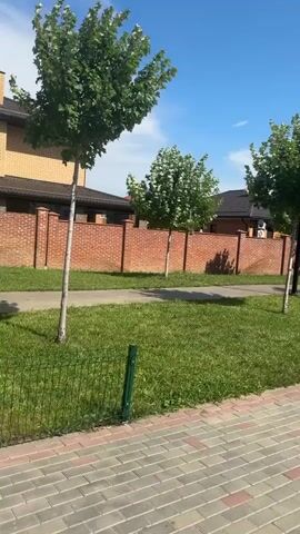 The video pans across a suburban street scene in Krasnodarskiy, Russia. The foreground features a patterned brick walkway with a well-maintained lawn and a short metal fence on its left. Several young, slender trees with lush green foliage are planted along the edge of the lawn, casting shadows on the grass. Behind the trees, a decorative brick wall separates the street from a row of houses and buildings. The houses have brick exteriors and darker roofs, and one has an air conditioning unit visible on its exterior wall. In the distance, taller apartment buildings and a smokestack with red and white markings are visible against a bright blue sky dotted with white clouds. Two figures, a woman and a child, are walking away from the camera on the distant sidewalk. The scene appears to be set on a sunny day, likely in the late morning or early afternoon, given the clear sky and the strong shadows. The overall impression is one of a peaceful, developed residential area. No visible text is present in the video.