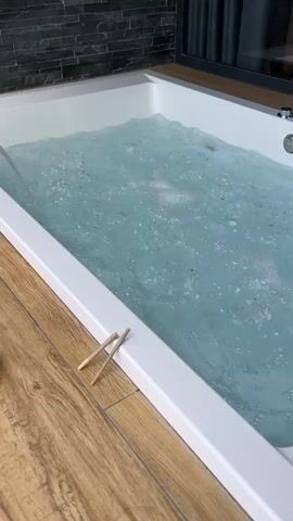 A close-up shot shows a bathtub filled with clear, bubbly water. Two chopsticks are placed on the white rim of the tub. The water is being agitated, suggesting it is a jacuzzi or hot tub. The background features a dark tiled wall and a wooden floor.