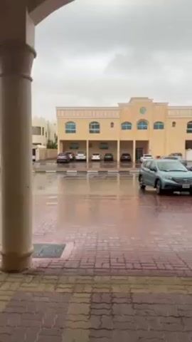 The video shows a rainy day scene from the perspective of someone standing under a covered area, likely a porch.  A brick-paved area in front of the person is wet and reflects light.  Across the paved area is a two-story, light-yellow building.  The ground floor of the building is essentially a series of covered parking spaces, each occupied by a vehicle.  From left to right, these cars appear to be a dark gray sedan, a white SUV, a white sedan, a black sedan, and a dark gray sedan.  To the right of the building, a blue-gray SUV is parked along the curb.  The sky is overcast and gray, suggesting a cloudy day.  There is no visible human activity.