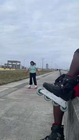 A person wearing black pants, a light blue t-shirt, and roller skates is roller skating on a paved path outdoors. Another person is partially visible in the foreground, also wearing roller skates and black pants. The background shows a grassy area, some buildings under construction, and a few tall buildings in the distance under a cloudy sky. The roller-skating person is moving away from the foreground person.