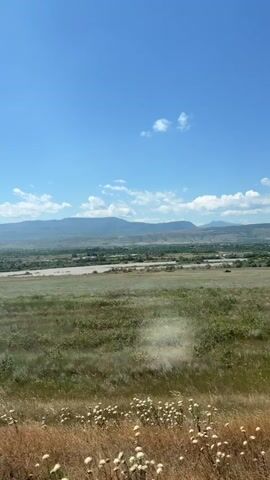 The video shows a panoramic view of a rural landscape under a clear blue sky with scattered white clouds. In the foreground, there is a field of tall green and brown grass, with some dried seed heads. In the middle ground, a wide, muddy river flows through a flat, treeless plain. In the background, rolling hills and mountains recede into the distance. The scene appears to be captured from a moving vehicle, possibly a train or car, as evidenced by a vertical pole on the right side of the frame and a slight blur that suggests motion. The overall impression is one of a vast, natural landscape on a sunny day.