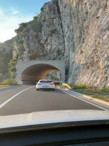 This video captures a scenic drive through a mountain tunnel in Brajici, Montenegro. The primary subject is a white car, seen from the perspective of another vehicle following closely behind. The car is traveling on a paved road that leads into a large, concrete tunnel carved into a steep, rocky mountainside.

The scene is set during the late afternoon or early evening, as indicated by the warm, golden sunlight illuminating the landscape and casting long shadows. The weather appears clear and pleasant, with a bright sky visible at the tunnel's entrance. The sheer rock face of the mountain dominates the background, showcasing its rugged texture and dramatic contours.

As the car enters the tunnel, the lighting shifts, with artificial lights within the tunnel illuminating the arched ceiling and walls. The tunnel is lined with red and white striped markings on the outside, and red graffiti is visible on the interior walls. The road inside the tunnel is asphalt with a clear white lane divider. After exiting the tunnel, the road continues to wind along the mountainside, offering expansive views of the rocky terrain and the sky.

The overall mood is one of travel and exploration, with a sense of awe inspired by the natural beauty of the mountainous landscape. The video captures the transition from the bright, open exterior to the enclosed tunnel and back out to the open road, highlighting the dramatic changes in light and scenery.

There are no discernible people visible besides those implied to be in the vehicles. The prominent objects are the two cars, the tunnel structure, the road, and the natural rock formations. The location cues, such as the mountainous terrain and the tunnel, strongly suggest a location like Brajici, Montenegro, known for its dramatic coastal roads and tunnels.

No significant text is clearly legible in the video, other than the striped markings and graffiti on the tunnel.