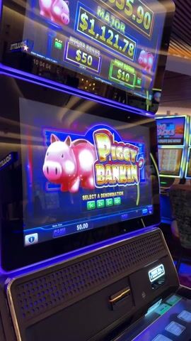 The video shows a close-up view of a "Piggy Bankin'" slot machine in a casino. The machine is brightly lit with blue and purple lights. The top screen displays the progressive jackpot amounts: "Major" at $1,121.78, "Minor Bonus" at $50.00, and "Mini Bonus" at $10.00. The main screen shows the "Piggy Bankin'" logo with a cartoon pig. The game prompts the user to "Select a Denomination," with options of 1c, 2c, 5c, and 10c. The machine's cash balance reads $0.00. Other slot machines are visible in the background, along with a casino chair. There is no person visible in the frame.