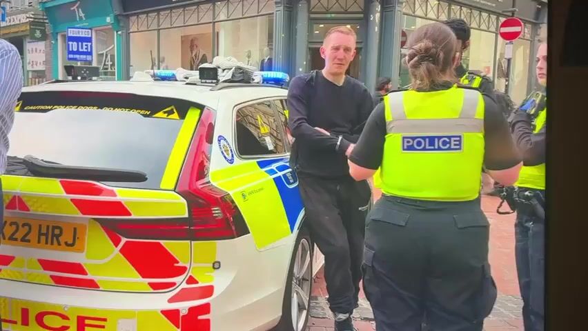 The video shows a street scene with a police car and several people. A man in handcuffs is standing next to the police car with two police officers. One officer is wearing a yellow vest with the word "POLICE" on the back. Another officer is standing further away. A man in a blue and white striped shirt is on the phone. In the background, there is a building with shops and a construction site with temporary fencing. The ground is paved with bricks. There are signs on the buildings and fences.