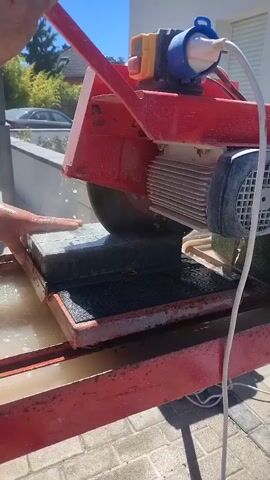 A person uses a wet tile saw to cut a dark gray rectangular object. The saw is red and has a blue electrical plug connected to it. The object being cut is placed on a metal platform and water is spraying around the saw blade as it cuts. The background shows a white building and a paved walkway.
