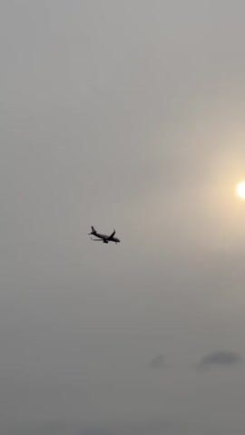 A drone shot shows an airplane flying through a hazy sky. The sun is visible as a bright circle in the upper right corner of the frame. The airplane, silhouetted against the sky, is in the center left of the frame. The airplane appears to be in flight and possibly preparing to land, as its landing gear is down. There are faint clouds in the lower part of the frame. The overall atmosphere is overcast and muted.