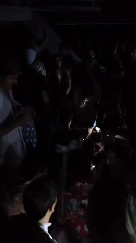 A crowd of people are dancing in a dimly lit space. Some are holding drinks and wearing sunglasses. One person raises their fist in the air, and another appears to be getting married with a floral crown. Most of the people are facing towards the camera, and the background is dark with a hint of white structure at the top.