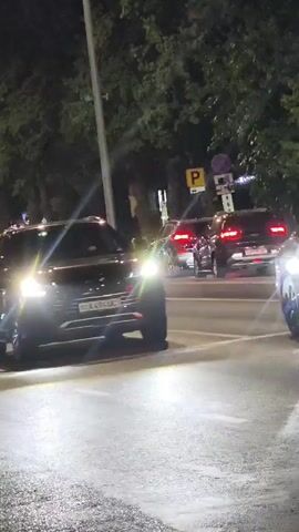 A black SUV drives down a street at night. The street is wet, reflecting the headlights of the car and streetlights. Trees line the street, and there are other cars and streetlights in the background. The SUV's headlights and taillights are illuminated. A license plate is visible on the back of the SUV.