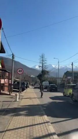 The video shows a street scene with snow-capped mountains in the background under a clear blue sky. The camera is stationary, capturing a view down a road lined with buildings and parked cars. A speed limit sign of 40 is visible.  A man walks across the street.  Trees and power lines are present. The sidewalk is made of patterned paving stones.
