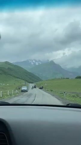 The video shows a view from inside a car, looking out at a winding road through a mountainous landscape under a cloudy sky. Several cars are parked or driving on the road. Two individuals are walking on the road in the distance. The surrounding hills are covered in green grass, with some patches of snow visible on the higher mountain peaks.