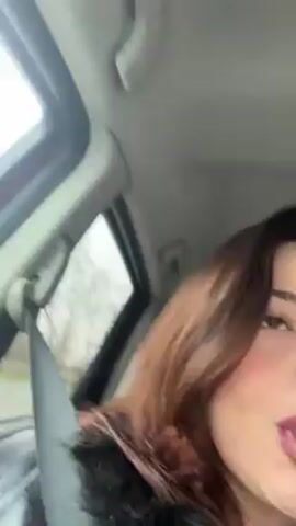 A young woman with shoulder-length brown hair is sitting in a car, wearing a black leather jacket and a gray hooded sweatshirt. She is wearing a seatbelt. The woman is looking at the camera and singing along to music. She seems happy and energetic. A red object can be seen in the background, possibly a car seat. Another person is sitting next to her. The car is in motion. The video is recorded from a close-up perspective inside the car.