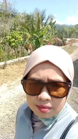 A medium shot captures a woman wearing a light pink hijab and large, dark-lensed sunglasses. She has on a light blue jacket and is looking directly at the camera. Her facial expression is neutral. The background consists of a dirt road with small stones and dry grass on the left, and lush green foliage, including palm trees and banana plants, on the right. The scene is illuminated by bright daylight, suggesting an outdoor setting, possibly in a tropical or subtropical region. The video appears to be a selfie.