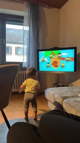 A young child stands on a wooden floor facing a television screen that displays an animation of bees. The child is wearing a light-colored t-shirt and gray pants, and is barefoot. To the left of the child is a dark-colored chair. To the right of the child is a light-colored sofa. A window with gray curtains is visible behind the child. The room appears to be a living room.