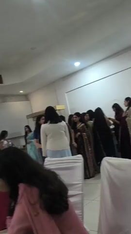 The video opens with a wide shot of a gathering of women in what appears to be an indoor event space. Several women are dressed in traditional Indian attire, specifically sarees, suggesting a formal or celebratory occasion. The lighting is a mix of overhead fluorescent lights and possibly some natural light from windows, creating a somewhat dim environment.

As the video progresses, the camera pans across the room, focusing on different groups of people. In one section, a group of women are clustered together, some appearing to be in conversation or interacting closely. One woman in a cream-colored top and light blue skirt is facing away from the camera, while another woman in a dark saree is visible behind her. The focus then shifts to a stage area decorated with a balloon arch made of blue and white balloons, and silver streamers. A woman in a red outfit is standing on the stage, seemingly addressing the attendees.

The video also includes shots of the audience, with many individuals seated and facing the stage. The seating appears to be a mix of chairs covered with white fabric. There are glimpses of other people, some with their backs to the camera, indicating a larger gathering.

Towards the end of the video, the camera briefly focuses on a smartphone with a decorative case, held by someone in the foreground. The background shows a continuation of the event space, with more attendees and decorative elements. The overall impression is that of a lively event, possibly a party, graduation, or cultural celebration, with a focus on the women attending and a designated stage area for a performance or speech.