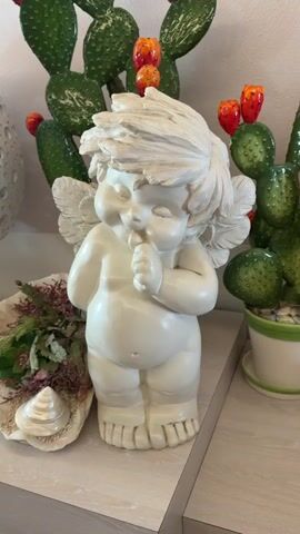 A white cherub statue is displayed on a shelf. The statue is of a cherub with closed eyes, its hands clasped to its mouth. Behind the statue are two prickly pear cacti with red fruit. To the left of the cherub is a shell with some dried foliage. The background is a light-colored wall.