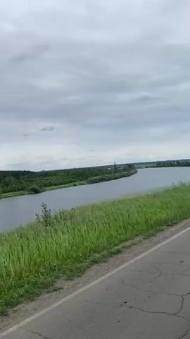 A POV video shows a view from a moving vehicle traveling along a paved road. To the left of the road, a wide, calm river flows, bordered by lush green grass. The opposite bank of the river is lined with dense green trees, extending into the distance under a cloudy sky. The road is cracked and weathered.
