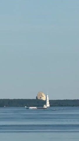 A distant shot shows a lighthouse on a body of water. A barge travels in front of it, and a large, spherical object is visible behind the lighthouse. The sky is clear and blue.