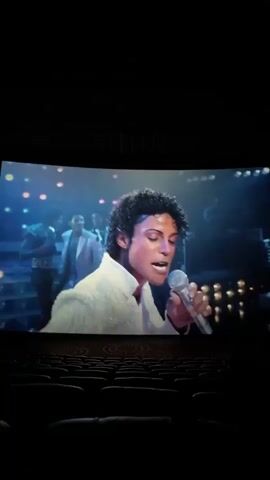 The video shows a movie screen in a dark movie theater. The screen displays Michael Jackson in concert, singing into a microphone while wearing a sparkly white jacket and black and white striped pants. Stage lights and silhouettes of band members are visible in the background. The theater is empty except for the person filming.