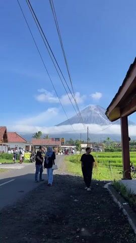 A street scene unfolds with a majestic volcano dominating the background, its peak shrouded in clouds. The sky is a vibrant blue with scattered white clouds. A paved road runs horizontally across the lower portion of the frame. To the left, a few buildings with red roofs are visible, along with a parked motorcycle. Several people are walking on the road and sidewalk. In the foreground, two individuals stand on a dirt path. One person, wearing a headscarf and jeans, walks towards the left, while another person, dressed in a black t-shirt and pants, stands facing the right. A lush green rice paddy stretches to the right of the road, with a few trees interspersed. Overhead, several power lines crisscross the frame. The overall atmosphere suggests a rural, possibly touristy, location.