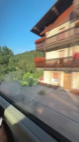 The video captures a view from a moving vehicle, likely a train, looking out of a window. The scene is dominated by lush green landscapes, with rolling hills covered in dense trees and patches of bright green grass. In the distance, a mountain range can be seen with a clear blue sky overhead, dotted with a few white clouds. The trees on the hills are a mix of deciduous and coniferous types, with some taller evergreen trees interspersed. The sunlight casts shadows across the grassy areas, indicating it is daytime. The movement of the vehicle causes a slight blur in the foreground and some of the closer trees, creating a sense of speed and motion. A building, possibly a house or a lodge, is visible nestled amongst the trees on one of the hillsides. The overall impression is of a scenic journey through a mountainous, forested region on a clear day.