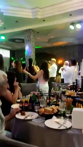 The video captures a lively indoor gathering, possibly a banquet or celebration, with a band performing on a raised platform in the background. The band, consisting of at least three men and two women, is seen singing and playing music, illuminated by colorful stage lights. In the foreground, several tables are laden with food and drinks, suggesting a meal is in progress. People are seated at the tables, some engaged in conversation while others focus on the performance. The overall atmosphere appears festive and energetic, with guests enjoying the music and company. The lighting is dim, with bright spotlights illuminating the performance area.