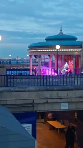 A video shows a band performing on an outdoor stage with purple and pink lights. The stage is located in a circular building with a blue roof, supported by columns. A blue railing runs in front of the stage. Below the stage, there is a walkway with a person walking by a table and a trash can. The sky is overcast and the lighting suggests it is evening.