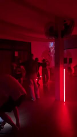 The video shows people dancing in a room lit with red light. Three people are at the center of the frame, moving to music that is playing. The background has a projection of snowflakes and a koi fish on a white wall. There is a person sitting in the lower left foreground, and fans are visible on the ceiling. Red light bars provide additional lighting. The dancers appear to be in a club or party setting.