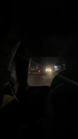 The video captures a nighttime view from inside a vehicle, looking out through the windshield. The scene is dark, illuminated by the headlights of passing vehicles and streetlights. The road appears wet, suggesting recent rain or damp conditions.

In the foreground, the interior of the car is mostly in shadow, with a portion of the steering wheel and dashboard visible on the left. A dark-colored object, possibly a blanket or clothing, is draped over the passenger seat on the right.

On the road ahead, several vehicles are visible. A motorcycle with a red taillight is moving away from the camera. Further down the road, headlights of other vehicles, including what appears to be a truck, are bright and slightly diffused due to the darkness and potentially some moisture in the air. One truck has "PUTRA" written on its side in white letters. There are also some buildings visible along the roadside, with lights on inside some of them. The overall impression is of a journey on a road at night in a populated area, possibly a town or village in Indonesia, given the location context. The atmosphere is subdued and the light conditions create a somewhat moody or contemplative feel. There are no clear indications of specific emotions, but the darkness and solitary perspective can evoke a sense of quiet travel.