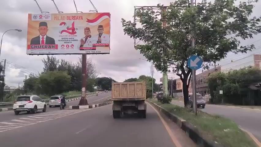 A car is driving on a highway. The sky is overcast. There is a truck in front of the car. There are other vehicles on the highway. A large sign with two men's faces on it is on the left side of the road. There are trees on both sides of the road. There are buildings on the right side of the road. The road has a median.