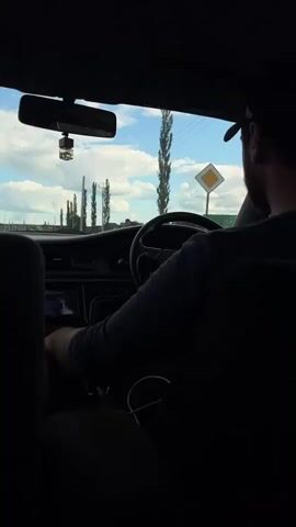 This is a shot from inside a car, looking forward through the windshield. The car is moving, as indicated by the blurred background and the timestamp. The driver, a man wearing a dark blue shirt and a baseball cap, is at the wheel, his right hand resting on the steering wheel. A rear-view mirror hangs from the top of the windshield, and a small, dark object, possibly an air freshener, dangles from it. The dashboard is visible, including a car stereo. Outside, the sky is blue with white clouds. Several tall, thin trees are visible, as well as what appear to be power lines and utility poles. In the distance, a person is walking. The interior of the car is dark, with the back seats mostly silhouetted.