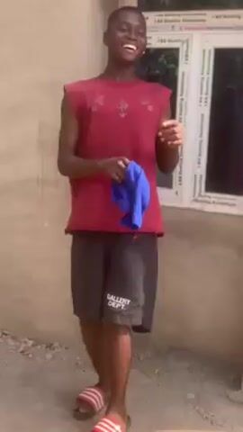 A young man stands outside a building. He is wearing a sleeveless maroon shirt, black shorts with “GALLERY DEPT.” printed on them, and red and white striped sandals. He is smiling and holding a blue cloth in his hands, gesturing with it. The building has a window with white framing. The ground is bare.