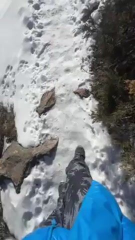 The video shows a first-person perspective of someone hiking on a snowy trail, likely in a mountainous region. The ground is covered in snow with visible footprints. The hiker is wearing dark camouflage patterned pants and a blue jacket. The sun is shining brightly in a clear blue sky. The landscape includes snow-covered mountains, trees, and rocks. As the video progresses, the view opens up to reveal a wider mountain vista.