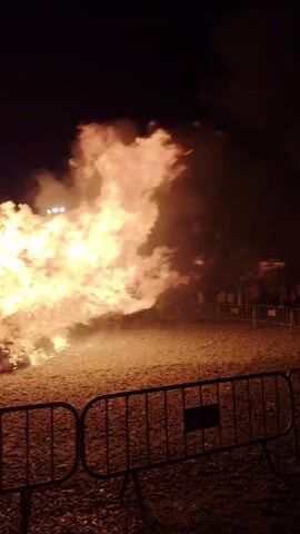 A large bonfire burns brightly at night, with flames reaching high into the dark sky. The fire is contained within a fenced-off area, and a crowd of people stands behind the barrier, observing the spectacle. The ground around the bonfire appears to be covered in gravel or dirt. In the background, trees and streetlights are visible, suggesting an outdoor urban or park setting. The smoke from the fire billows upwards, obscuring some of the background details.