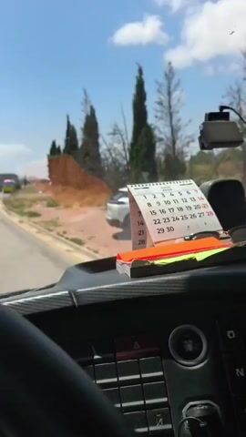 The video provides a first-person perspective from inside a moving vehicle, likely a bus or truck, during daytime. On the dashboard, a calendar displaying "JULY 2024" is visible, along with sunglasses and a stack of colored notes. A dashboard camera is mounted on the windshield, and a sticker reads "V-SET=90 KM/H".

The outdoor scene unfolds under a bright blue sky with scattered clouds. The landscape consists of dry, yellowish-brown grass and dirt areas, interspersed with tall, slender green trees, resembling cypresses. The vehicle is driving on a paved road with distinct yellow and red curb markings.

As the vehicle progresses, it reveals a white car parked or veered off the road into the dry, grassy terrain, with its driver's side door visibly open. Nearby, partially obscured by trees, an emergency vehicle, possibly a fire truck, is seen with flashing lights, suggesting an incident or accident has occurred. In the background, a power line tower is also visible. The overall setting is consistent with a dry, open area, possibly near an industrial zone given the provided location context of Polígon Industrial Dels Vents, Spain.