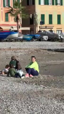 The video shows a scenic view of a beach in a European town with colorful buildings. The beach is covered with pebbles, and there is a small pool of water near the shore. In the first shot, two people are sitting on the beach. There are several small boats parked near the shore, some on trailers. The buildings behind the beach are painted in different pastel colors. The sky is blue with some clouds. The video then zooms out to show more of the beach and buildings. A wooden dock extends into the water. Sirens can be heard in the background.