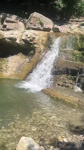 A narrow waterfall cascades down a rocky cliff face into a pool of water. The rocks are layered and appear to be sedimentary. There are some green plants growing on the rocks. On the top of the cliff, there are some red and white objects, possibly towels or clothing, spread out on the rocks. The water is clear and the bottom of the pool is visible, showing rocks and pebbles. The sun is shining, creating highlights on the water and rocks.