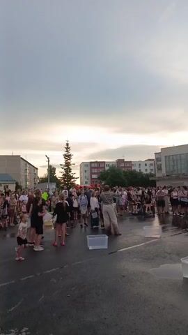 A group of children are participating in an outdoor activity in front of a crowd of onlookers. The activity appears to involve a relay race where children run and pour water from buckets into larger containers. The ground is wet, suggesting recent rain or a water-based activity. The sky is overcast with some hints of sunset colors. Buildings are visible in the background, indicating an urban or suburban setting.