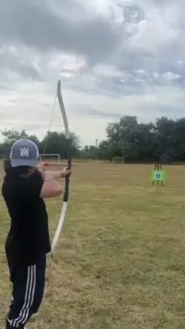 A medium shot captures a person aiming an arrow with a bow at a target in a field. The person, wearing a dark t-shirt, dark sweatpants with white stripes, and a baseball cap, is seen from the back, drawing the bowstring back. The bow itself is light in color with dark accents. In the background, the field is green with two soccer goals visible. Further back, there are trees. The target is visible in the center of the frame, brightly colored with concentric circles. The sky is overcast with cloudy patterns. The arrow is released and travels towards the target.