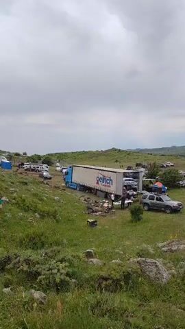 The video shows a large outdoor gathering in a grassy, hilly area with scattered rocks. The sky is overcast with clouds. A semi-truck with a trailer labeled "Goinch" is parked with its rear doors open, and several vehicles are positioned around it. Many other vehicles are parked throughout the scene, including SUVs, sedans, and trucks. There are several white tents set up in the distance, and a large crane is visible in the background. Some people are standing near the vehicles and tents, and a few individuals are interacting. The overall impression is of a large event or festival being held outdoors, possibly a car show or a gathering with vendors.