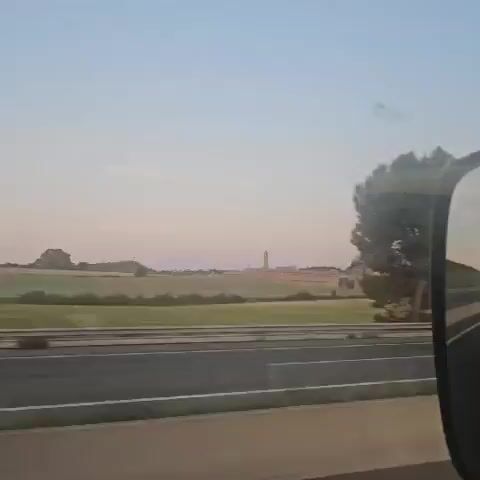 The video shows a view from a moving vehicle, likely a train or car. The camera pans past a tree, revealing a landscape with a field and a distant building with a tall spire or tower. The sky is a pale pink or purple, suggesting either dawn or dusk. The focus is blurry, particularly at the end of the video when the camera zooms in on the distant building.